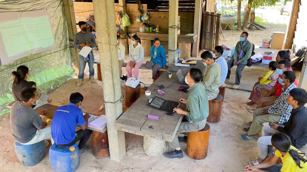 A group of people in Cambodia sit underneath a structure at tables with laptops and notebooks