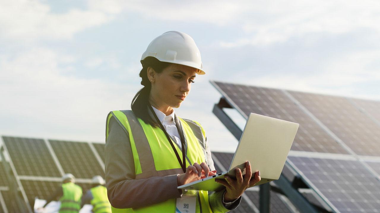 Female engineer in safety gear using laptop with solar panels in the background