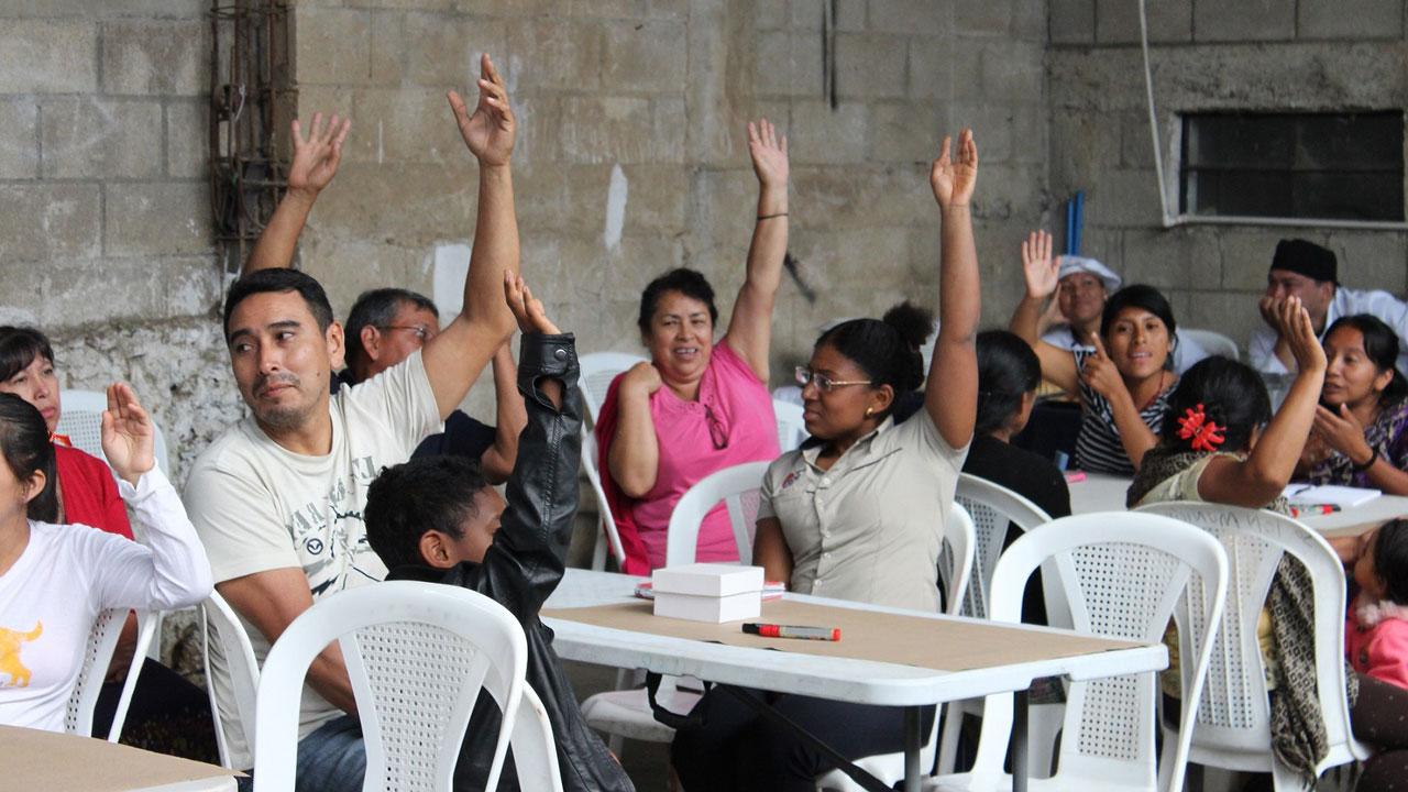 A group of people sitting around tables indicates their vote with their raised hands